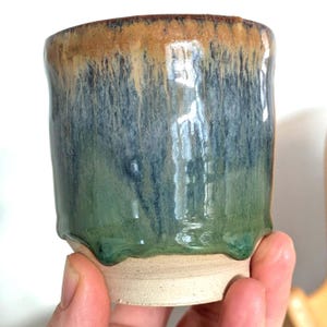 May include: A handcrafted ceramic cup with a distinctive glaze. The rim is a warm brown, fading into a deep blue and then a vibrant green at the base. The cup features a textured, dripped glaze effect and an unglazed base.