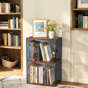 May include: A two-tiered, dark brown bookshelf with black mesh sides, filled with books. A framed painting and a vase of flowers sit on top. Additional wooden bookshelves are visible in the background, along with a woven basket.