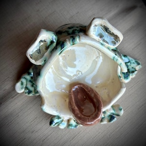 May include: A ceramic frog-shaped dish with a light beige interior and a brown, oval-shaped center. The exterior features green and blue speckled accents. The dish has raised eyes and leaf-like details around the edges.