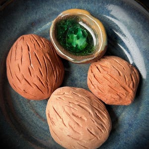 Puede incluir: Colección de piezas de cerámica hechas a mano sobre un plato azul. Las piezas incluyen tres objetos texturizados, marrones, con forma de nuez y un objeto pequeño, abierto, en forma de cuenco con un centro verde y un borde dorado.