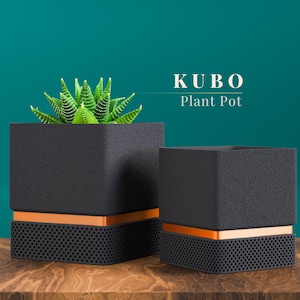 May include: Two black square plant pots with orange accents and a perforated base sit on a wooden surface. One pot contains a green succulent plant. The text "KUBO Plant Pot" is displayed above the pots.