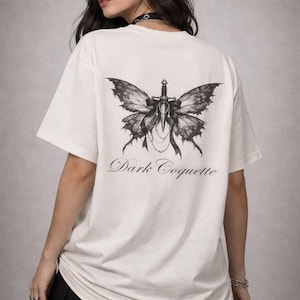 Peut inclure: T-shirt blanc cassé avec un motif de papillon noir et gris orné d'une épée. Les ailes du papillon sont finement détaillées, et le texte "Dark Coquette" est écrit en dessous en écriture cursive.