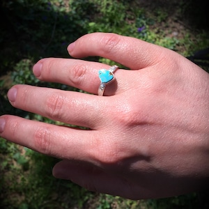 Könnte beinhalten: Ein silberner Ring mit einem herzförmigen türkisfarbenen Stein. Der Ring befindet sich an einer Hand mit einem verschwommenen grünen Hintergrund. Das Band des Rings hat ein strukturiertes Design.
