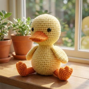 Op de afbeelding: Een handgemaakt, gehaakt eendenspeelgoed. De eend is voornamelijk geel met een oranje snavel en poten. Het heeft zwarte knoopogen en zit op een houten oppervlak. Op de achtergrond zijn potplanten te zien.
