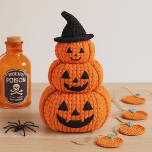 Könnte beinhalten: Gestapelte, gehäkelte Kürbisdekoration mit schwarzem Hexenhut. Die orangefarbenen Kürbisse haben lächelnde Jack-o'-Lantern-Gesichter. Eine Flasche mit der Aufschrift "Witch's Poison" und eine schwarze Spinne sind ebenfalls vorhanden, zusammen mit einer Kürbisgirlande, alles perfekt für Halloween.