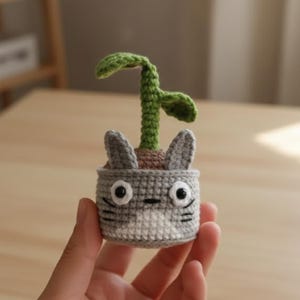 Może przedstawiać: Szydełkowy doniczka w kształcie postaci z kreskówki, z szarymi i białymi detalami, dużymi czarnymi oczami i zielonym pędem. Doniczka ma dwa szare uszy i małą zieloną roślinę wyrastającą z góry.