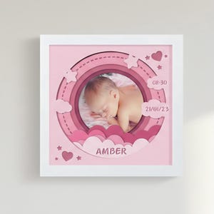 Puede incluir: Obra de arte en papel enmarcada en rosa con un bebé durmiendo en el centro. El diseño incluye círculos en capas, nubes, estrellas y corazones. Se muestra el nombre "AMBER", junto con la fecha y la hora.