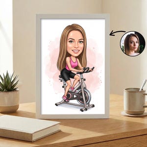 Könnte beinhalten: Gerahmte Cartoon-Illustration einer Frau auf einem Heimtrainer. Sie trägt ein rosa Oberteil und schwarze Shorts. Der Hintergrund hat einen rosa Aquarell-Effekt. Der Rahmen ist weiß, und das Bild befindet sich auf einer Holzoberfläche.