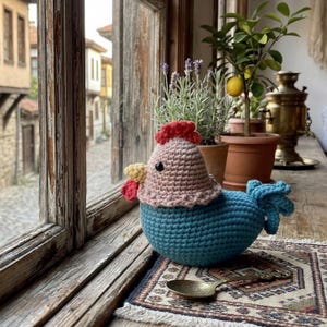 May include: A handcrafted crochet chicken in pink, blue, red, and yellow sits on a patterned rug near a window. The chicken has a red comb, yellow wattle, and a blue body. The background includes a view of a cobblestone street.