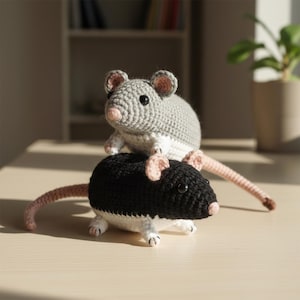 May include: Two crocheted toy rats. One is gray and white, the other is black and white. Both have pink ears, noses, and long tails. The toys are sitting on a light-colored surface.