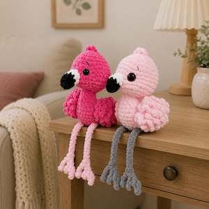 May include: Two crocheted flamingo plush toys sitting on a wooden surface. One flamingo is bright pink with light pink legs, while the other is light pink with gray legs. Both have black and white beaks and dark round eyes with stitched eyebrows.