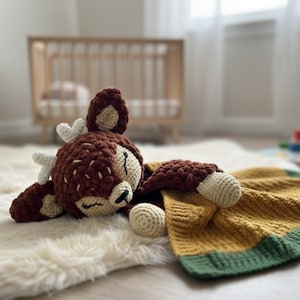 May include: A crocheted deer-shaped baby security blanket. The deer is brown with cream-colored accents and white antlers. The blanket is mustard yellow with a green border. The toy is lying on a white, fluffy rug.