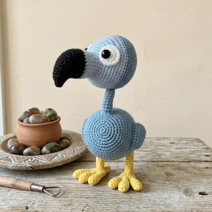 Könnte beinhalten: Ein handgefertigter blauer Häkelvogel mit schwarzem Schnabel, weißem Auge und gelben Füßen. Der Vogel steht auf einer Holzoberfläche. Ein kleiner Terrakottatopf mit Steinen und ein Holzwerkzeug befinden sich im Hintergrund.