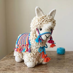 May include: A handmade crochet llama in cream with a colorful blanket and halter. The llama has a fluffy, textured body, small ears, and a detailed face. The blanket features stripes of blue, orange, pink, and yellow. Decorative tassels add a playful touch.