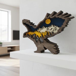 May include: A wooden eagle sculpture with a layered design. The sculpture features a mountain landscape with a yellow moon, white mountains, and a dark blue sky. The eagle is brown and stands on a white shelf.