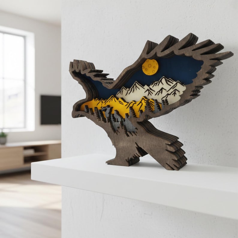 May include: A wooden eagle sculpture with a layered design. The sculpture features a mountain landscape with a yellow moon, white mountains, and a dark blue sky. The eagle is brown and stands on a white shelf.