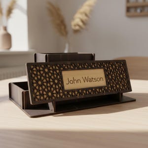 Op de afbeelding: Een donkerbruine houten bureau-organizer met een decoratief geometrisch patroon. De organizer heeft een rechthoekig naamplaatje met de tekst "John Watson". De bureau-organizer heeft meerdere compartimenten en een gelaagd ontwerp.