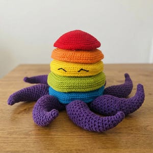 May include: A handmade crochet octopus toy with a rainbow-colored stack of rings on top. The rings are red, orange, yellow, green, and blue. The octopus has purple tentacles and a smiling face.