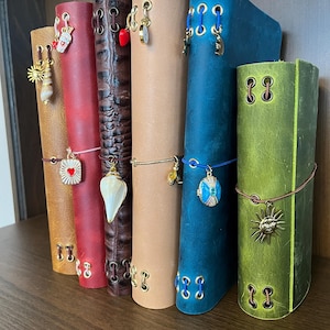 May include: A collection of leather journals in various colors, including tan, red, and green, arranged on a wooden shelf. Each journal is adorned with decorative charms and clasps, adding a personalized touch to the stationery.