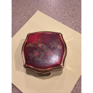 May include: A square-shaped metal compact case with rounded corners and a metallic gold-toned frame. The top surface features a deep red background decorated with a central illustration of a peacock surrounded by colorful floral accents and foliage.