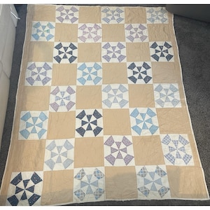May include: A rectangular quilt featuring a grid pattern of alternating solid tan squares and patterned squares. The patterned squares contain blue and white pinwheel designs in various shades of light blue, navy, and periwinkle with floral prints.