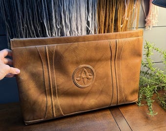 May include: A tan rectangular leather clutch bag featuring a centered circular embossed logo with the letter A. The bag has two vertical stitched panels on the front surface, a smooth texture, and a top zipper closure along the edge.