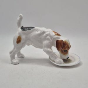 Può includere: Una statuetta in porcellana di un Jack Russell Terrier bianco con macchie marroni sulla testa e sul dorso, in posizione abbassata mentre mangia da un piccolo piattino bianco rotondo con un sottile bordo argentato, su sfondo bianco.