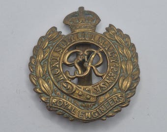 Op de afbeelding: Een vintage messing Royal Engineers pet-insigne. De badge heeft een kroon boven een cirkelvormig embleem met de letters "GR" en de woorden "ROYAL ENGINEERS" en "HONI SOIT QUI MAL Y PENSE" in een kransontwerp.