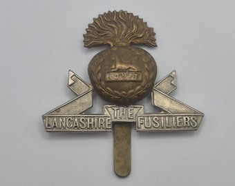 Op de afbeelding: Een metalen badge van de Lancashire Fusiliers. Het heeft een centraal embleem met een leeuw en een kroon, bekroond door een vlamachtig ontwerp. De tekst "Lancashire The Fusiliers" is zichtbaar. De kleuren zijn goud en zilver.