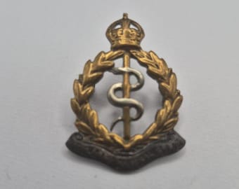 May include: A metallic military cap badge featuring a central vertical rod entwined with a silver-toned serpent, enclosed within a golden laurel wreath topped by a crown, with a dark, curved scroll base positioned at the bottom of the emblem.