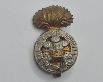 May include: A silver and gold Royal Welsh Fusiliers cap badge. The badge features a circular design with text and a central emblem of a crown and feathers, topped with a gold flame.