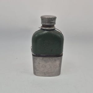 Op de afbeelding: Een kleine zakfles met een donkergroen, gestructureerd lederachtig bovendeel en een gladde, geborstelde zilverkleurige metalen onderkant. De fles is voorzien van een bijpassende zilverkleurige schroefdop en een dun donker koord rond de hals.