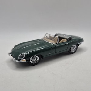 May include: A dark green die-cast model of a convertible sports car featuring a long bonnet, a beige interior, a clear windscreen, and silver wire-spoke wheels. The vehicle has a sleek, low-profile body design with chrome accents on the front bumper.