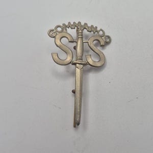 Puede incluir: Una insignia plateada del Comando SIS. La insignia presenta las letras "SIS" con una daga que las atraviesa. La palabra "COMMAND" está arqueada sobre las letras. La insignia es de metal.