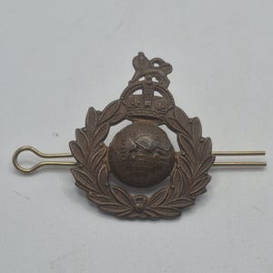 May include: A bronze military badge with a crown above a globe, surrounded by a laurel wreath. A pin is attached to the back for fastening. The design is intricate and likely vintage.