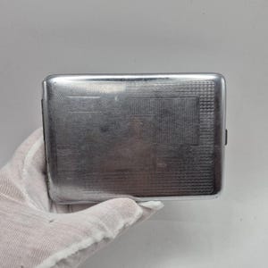 May include: A rectangular silver-toned metal cigarette case featuring a polished finish with a subtle, etched geometric pattern across the front surface. The case has rounded corners and a slim, flat profile, showcasing a metallic sheen.