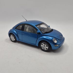 Pode incluir: Um modelo em escala azul metálico de um carro Volkswagen New Beetle com carroceria arredondada, rodas prateadas, uma pequena antena no teto e um interior visível com assentos claros, visto de um ângulo lateral elevado.