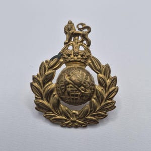 May include: A brass-colored metal military badge featuring a central globe encircled by a laurel wreath. Above the globe sits a crown topped with a standing lion figure. The design is embossed with intricate details throughout the metallic surface.