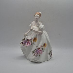 Puede incluir: Una figura de porcelana de una persona con cabello castaño claro recogido en un moño, vistiendo un vestido blanco de hombros descubiertos con mangas abullonadas. El vestido presenta un estampado floral con flores rosas, moradas y amarillas.