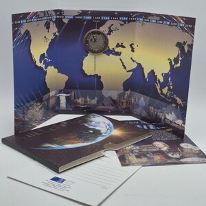 Puede incluir: Un set de moneda conmemorativa con una moneda plateada incrustada en una tarjeta de presentación de tres pliegues. La tarjeta muestra un mapa mundial con una escala horaria de 0100 a 2400, monumentos globales y una portada del Milenio.