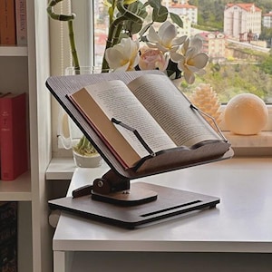 Könnte beinhalten: Ein hölzerner Buchständer mit einem geöffneten Buch. Der Ständer ist dunkelbraun mit einem rechteckigen Sockel und einer schrägen Oberseite, mit Metallstützen. Im Hintergrund sind Blumen und eine Stadtlandschaft zu sehen.