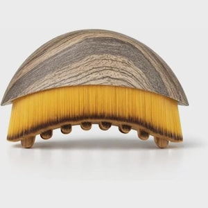 Può includere: Una spazzola in legno con una parte superiore a cupola e a strisce e setole gialle. La spazzola ha una forma curva con piccoli piedini arrotondati. Il legno ha un motivo a venature naturali e le setole sono fitte.