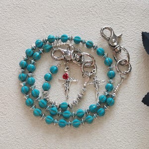 May include: A turquoise beaded rosary with silver-tone metal accents. The rosary features a cross pendant with a red rose and a lobster clasp. The beads are a vibrant turquoise color, and the design is circular.