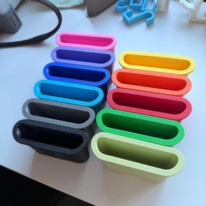May include: A collection of eleven small, rectangular, open-top containers arranged in two columns. The containers are made of solid-colored plastic in shades of pink, purple, dark blue, light blue, gray, black, yellow, orange, red, green, and lime.
