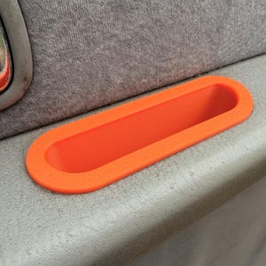 May include: An orange, oval-shaped car door handle insert. The insert is made of a textured material and is set against a gray interior. The handle insert is designed to fit into the door panel.