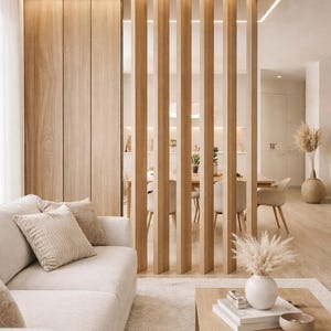 May include: A modern living room with a neutral color palette. A light-colored sofa with throw pillows sits on a plush rug. A wooden coffee table holds books and a vase with dried flowers. Vertical wooden slats create a room divider.