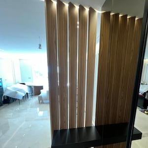 May include: Interior architectural detail featuring vertical wooden slats with integrated lighting. A black shelf is mounted on the wooden structure. The floor is tiled with a light-colored marble-like material, and the ceiling has recessed lighting.
