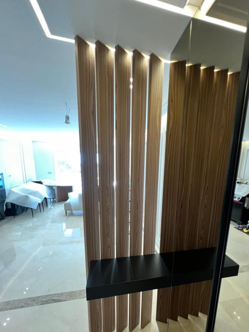 May include: Interior architectural detail featuring vertical wooden slats with integrated lighting. A black shelf is mounted on the wooden structure. The floor is tiled with a light-colored marble-like material, and the ceiling has recessed lighting.