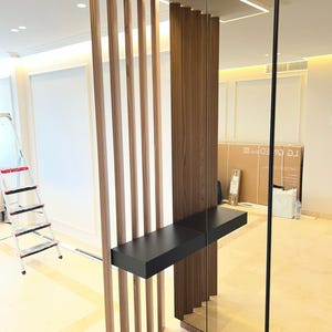May include: A modern room divider featuring vertical wooden slats and a dark wood panel. A black shelf is attached to the divider. A large mirror is on the right side of the divider. The room has a bright, contemporary design.