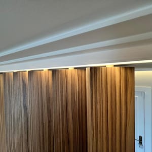 May include: A close-up of a wooden sliding door with vertical grain patterns. The door is a warm brown color with lighter streaks. The ceiling above has white beams and recessed lighting. The door is partially open, revealing a white door frame and wall.